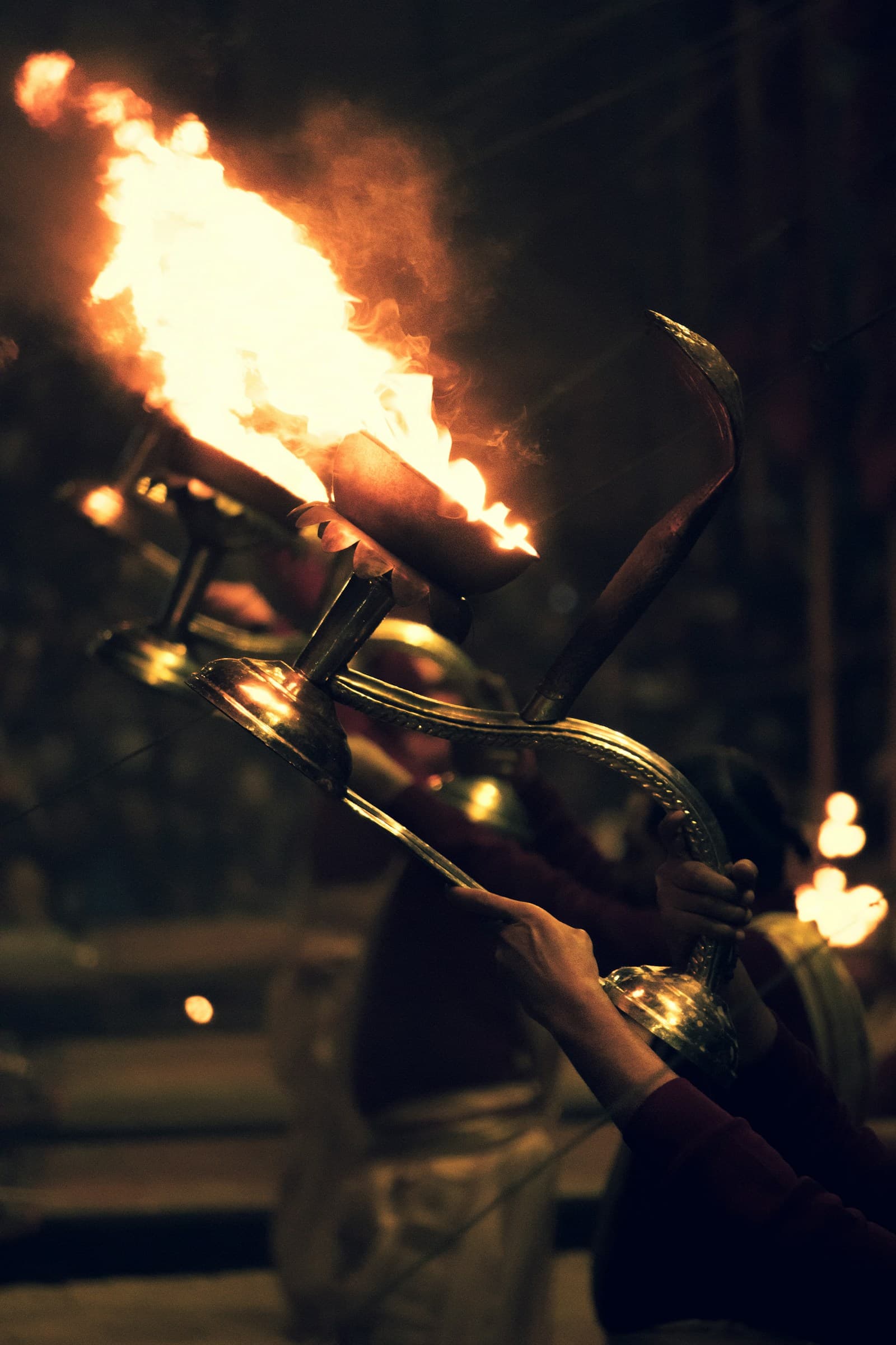 Priests holding multi-tiered brass aarti lamps with bright flames during an evening Ganga Aarti ceremony