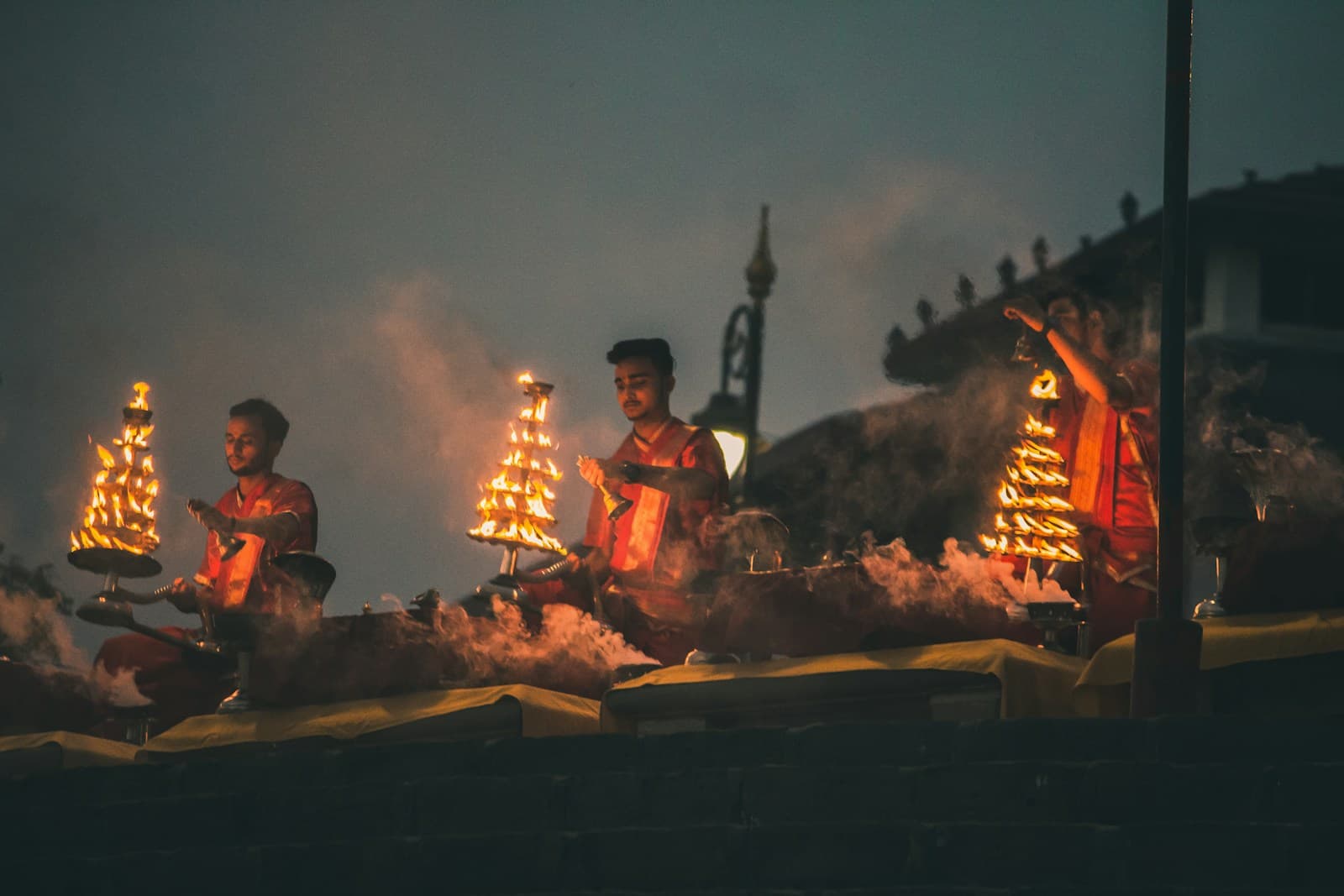 Priests in saffron robes performing evening Ganga Aarti with multi-tiered brass lamps on the ghats